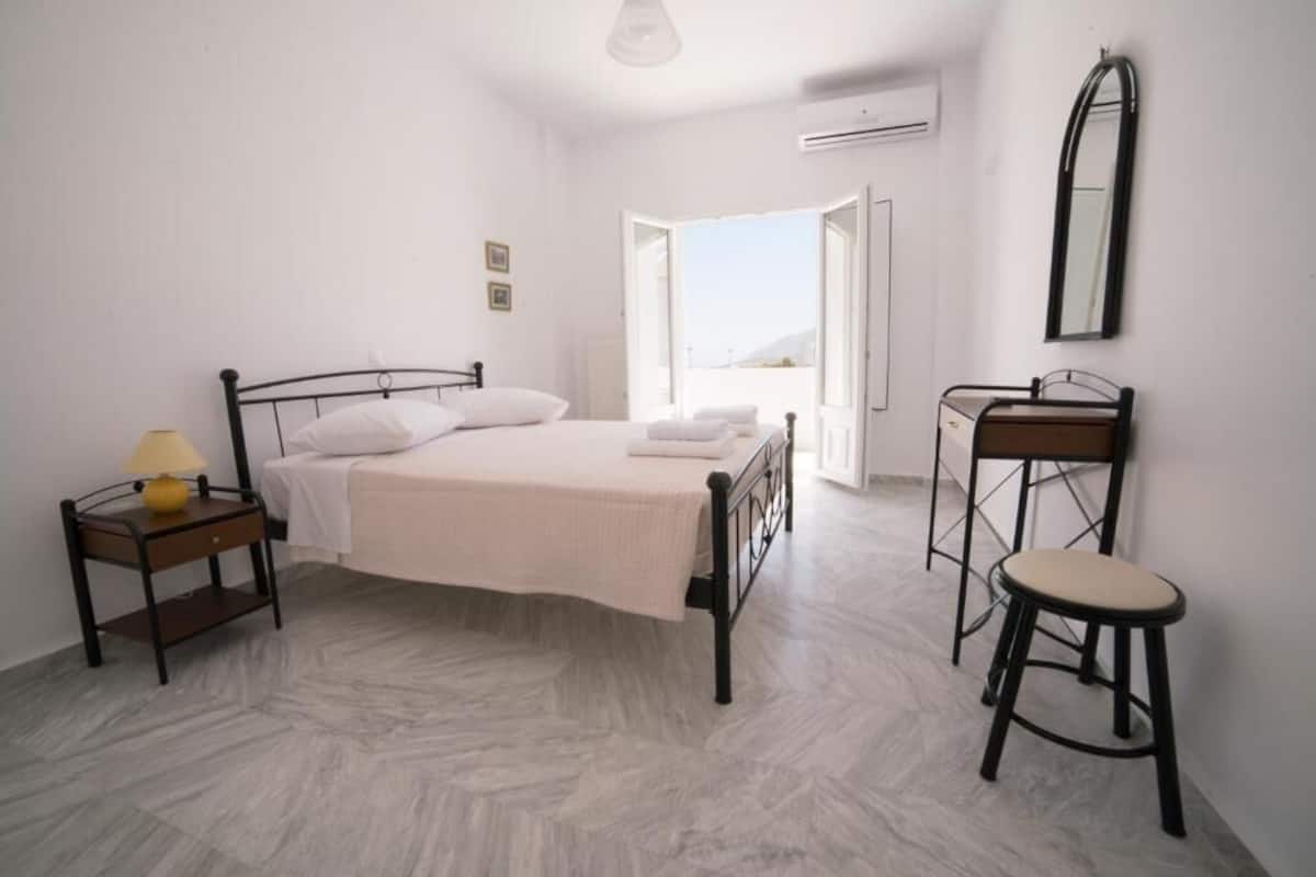 Un dormitorio con cama, mesita de noche, silla y vistas a través de una puerta abierta.