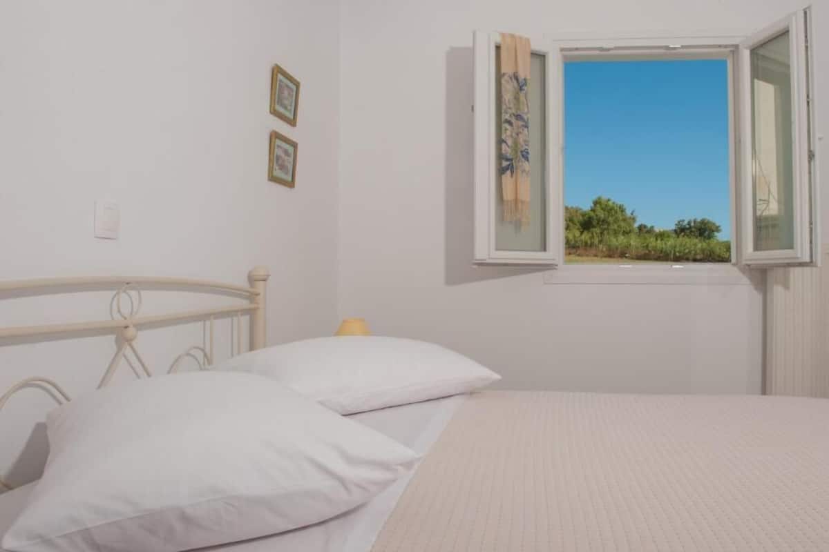 Un dormitorio con cama, ventana con vista a vegetación y pared con dos cuadros enmarcados.