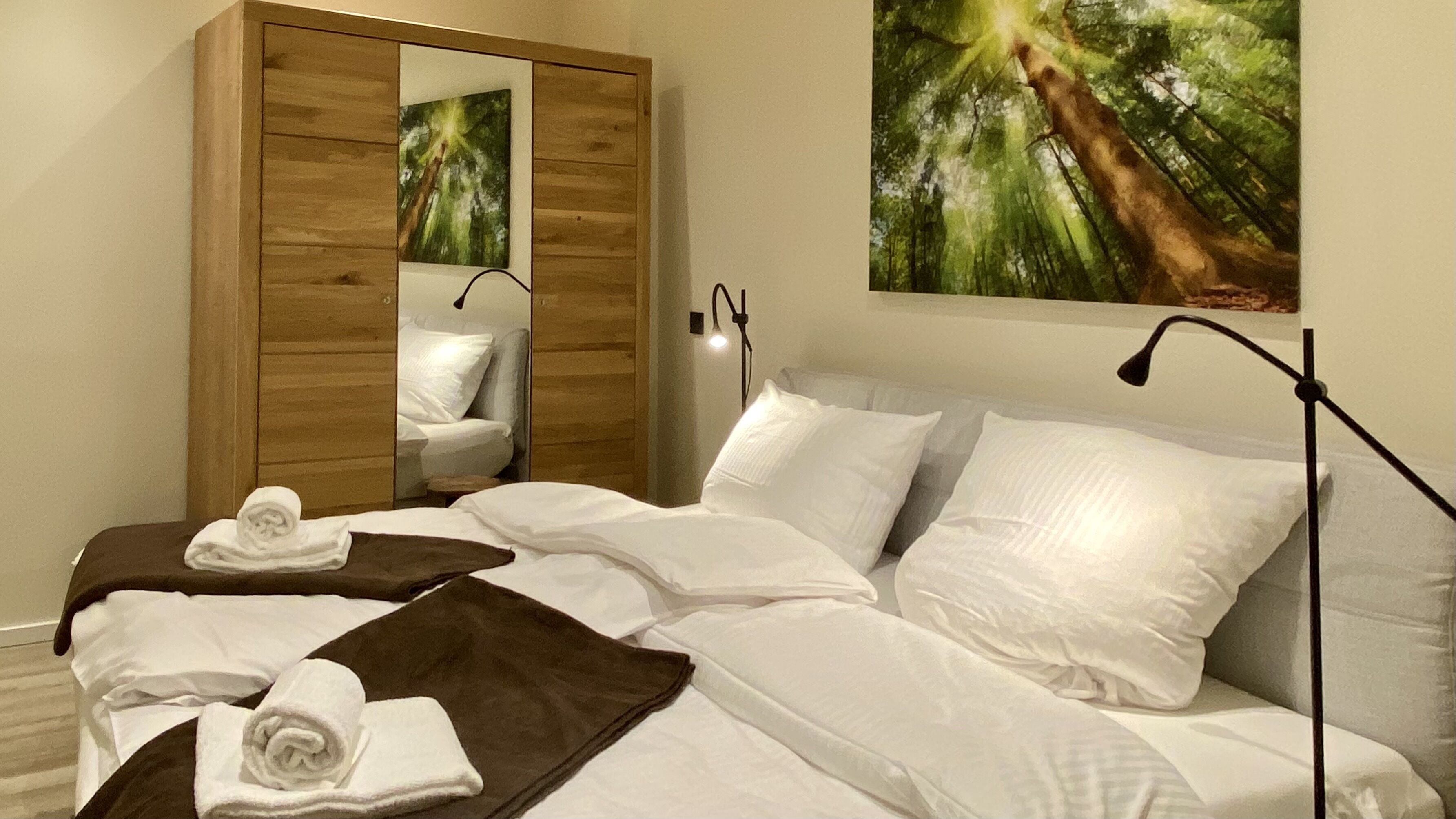 Habitación de hotel con una cama grande, dos almohadas, un armario de madera, un espejo y un cuadro de un bosque.