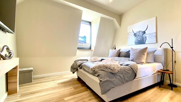 A modern bedroom with a large bed, a window, and a painting on the wall. - App. 4 - wohlBleben (4 Zi, 6 Pers.) (Braunlage)