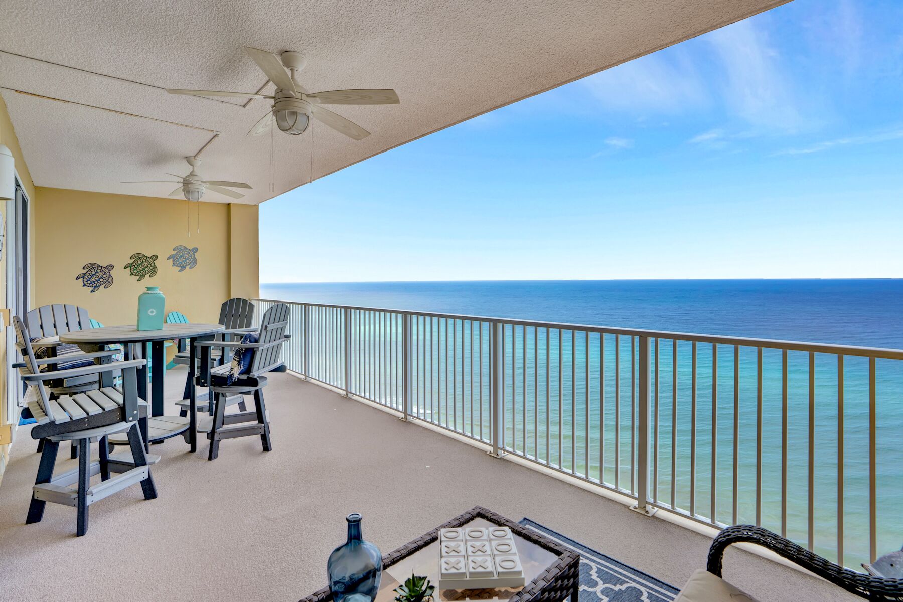 A balcony with a view of the ocean, featuring a table and chairs, a ceiling fan, and a decorative wall.