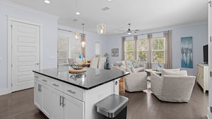 Una cocina moderna con armarios blancos, encimera negra e iluminación colgante. Hay una zona de comedor con un sofá blanco y un televisor.