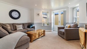 A living room with two sofas, a coffee table, a clock, and a window with blinds. - 3 South Beach Court - Cosy Seafront Apartment (Tenby)
