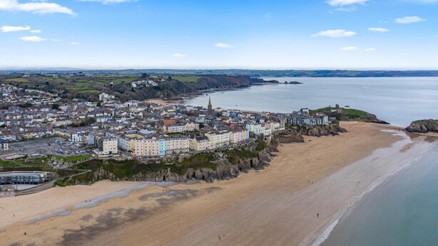 An aerial view of a coastal town with a sandy beach, a large body of water, and a dense cluster of buildings with varying architectural styles. - 3 South Beach Court - Cosy Seafront Apartment (Tenby)