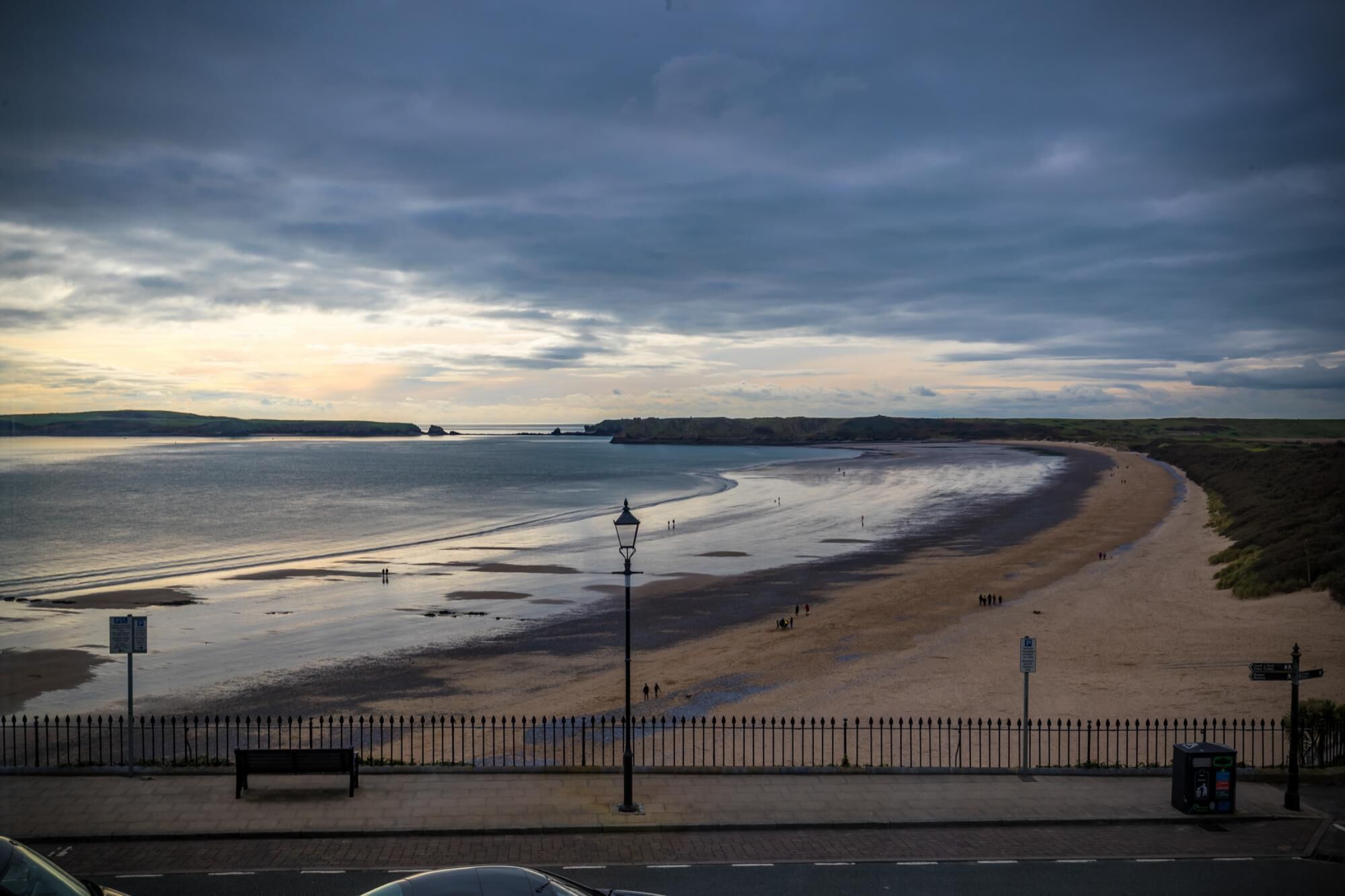 A beach with a curved shoreline, a bench, a lamp post, and a signpost.