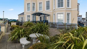 A multi-story building with a blue entrance canopy, stone planters, and a paved walkway. - 3 South Beach Court - Cosy Seafront Apartment (Tenby)
