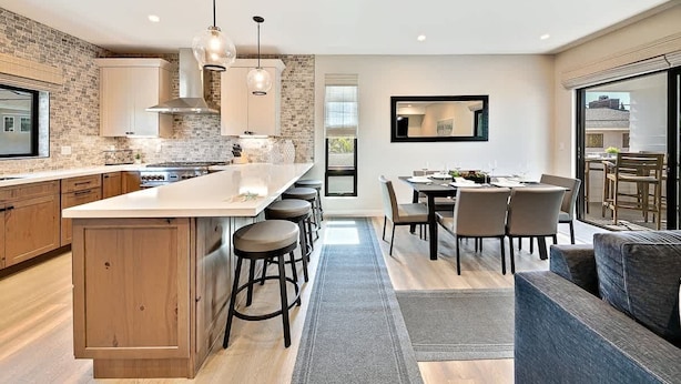 A modern kitchen with a central island, bar stools, a dining area with a table and chairs, and a large window.