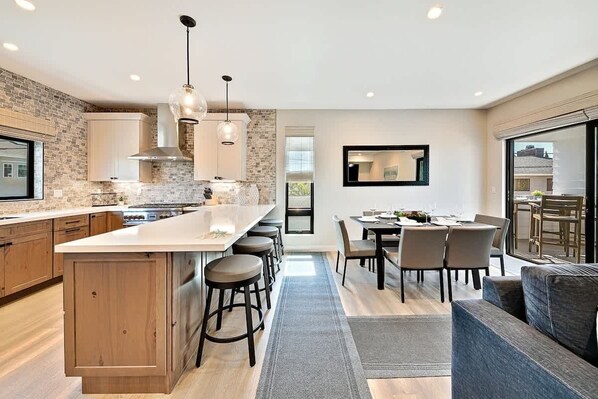 A modern kitchen with a central island, bar stools, a dining area with a table and chairs, and a large window. - Ocean Views - Jacuzzi - Rooftop Deck-walk to beach (San Diego)