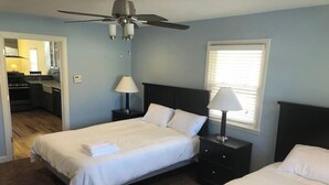 A bedroom with a bed, two nightstands, a ceiling fan, a window with blinds, and a view of a kitchen.