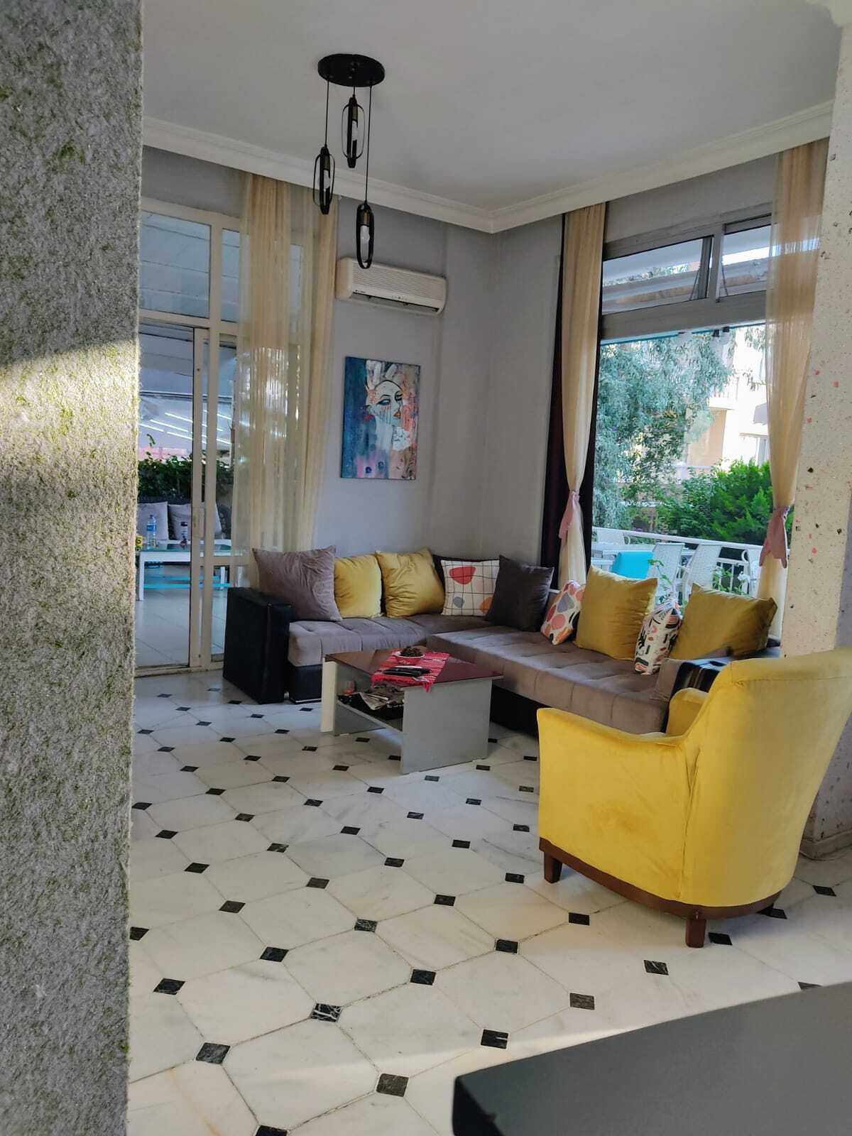 A modern living room with a grey sofa, yellow armchair, and a coffee table. There is a large window with curtains, an air conditioning unit on the wall, and a painting above the sofa.