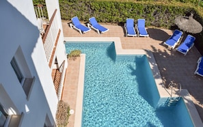 A rectangular swimming pool with clear blue water, surrounded by white walls and a tiled deck. There are multiple blue lounge chairs and a thatched umbrella.
