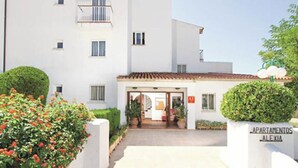 Un bâtiment blanc avec un toit recouvert de tuiles, une petite entrée dotée de portes vitrées et une enseigne indiquant « APARTAMENTOS ALEXIA ».