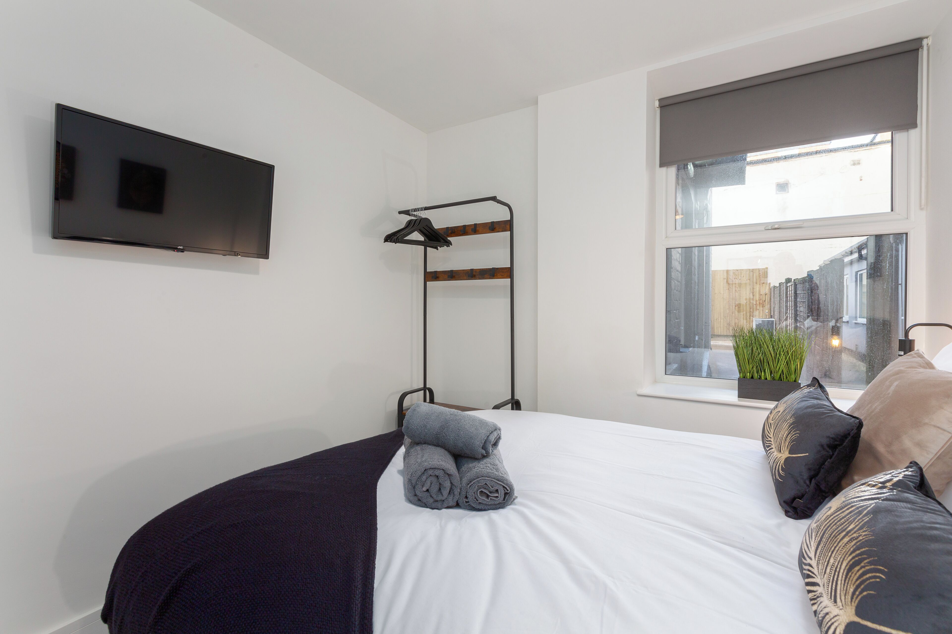 A neatly made bed with rolled towels, a mounted TV, a window with a view of the outside, and a plant on the windowsill.