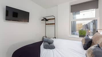 A neatly made bed with rolled towels, a mounted TV, a window with a view of the outside, and a plant on the windowsill.