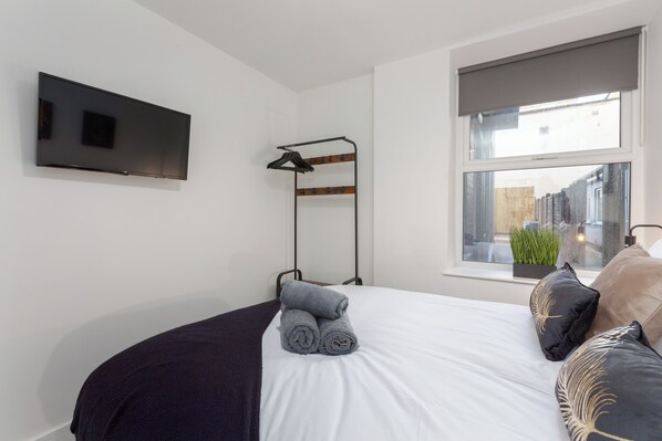 A neatly made bed with rolled towels, a mounted TV, a window with a view of the outside, and a plant on the windowsill.
