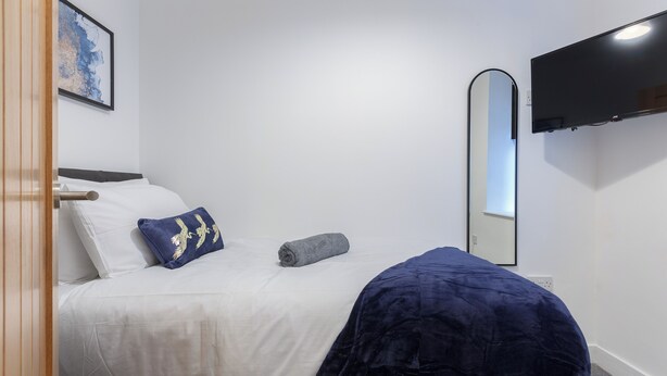 A bedroom with a bed, a blue pillow, a grey towel, a blue blanket, a wall-mounted TV, and a framed picture. - Charnley House Apartment 2 (Blackpool)