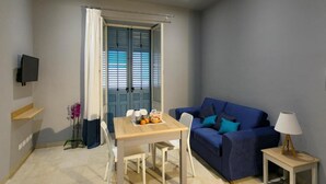 A compact living space with a dining table, a blue sofa, a small side table, and a TV mounted on the wall. - Superior Suite (Trapani)