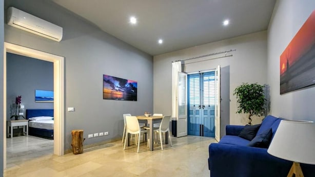 A modern living room with a blue sofa, a dining area with a table and chairs, a large window with blue shutters, and a painting on the wall. - Deluxe Suite (Trapani)