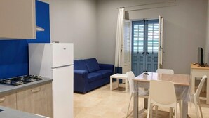 A compact kitchen with a gas stove, a white refrigerator, a blue sofa, a dining table with chairs, and a window with blinds. - Deluxe Suite (Trapani)