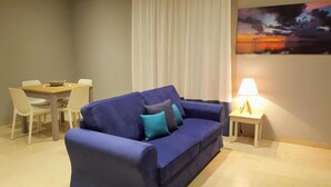 A room with a blue sofa, a small table with a lamp, and a dining area with chairs. - Deluxe Suite (Trapani)