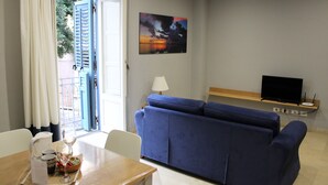 A living room with a blue sofa, a TV on a stand, a painting on the wall, and a dining area with a table and chairs. - Deluxe Suite (Trapani)