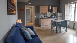 Una sala moderna con un sofá azul, una mesa de comedor con sillas, una cocina con armarios de madera y un refrigerador, y una puerta que da a otra habitación.