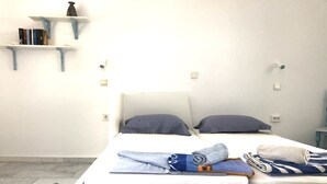 Une chambre avec un lit simple, une tête de lit blanche, deux oreillers, une couverture pliée et une petite table de chevet avec une lampe. On y trouve également des étagères murales avec des livres et un petit objet décoratif.