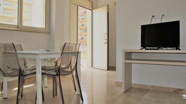 Een moderne kamer met een eettafel en stoelen, een televisie op een standaard, en een deur naar een balkon.