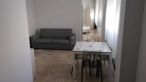 Een moderne, minimalistische woonruimte met een grijze bank, een eettafel met stoelen en een gang die naar een slaapkamer leidt.