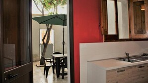 Une cuisine  équipée d’un évier et de placards, un coin repas avec une table et des chaises, et une vue sur un arbre à travers une porte ouverte.