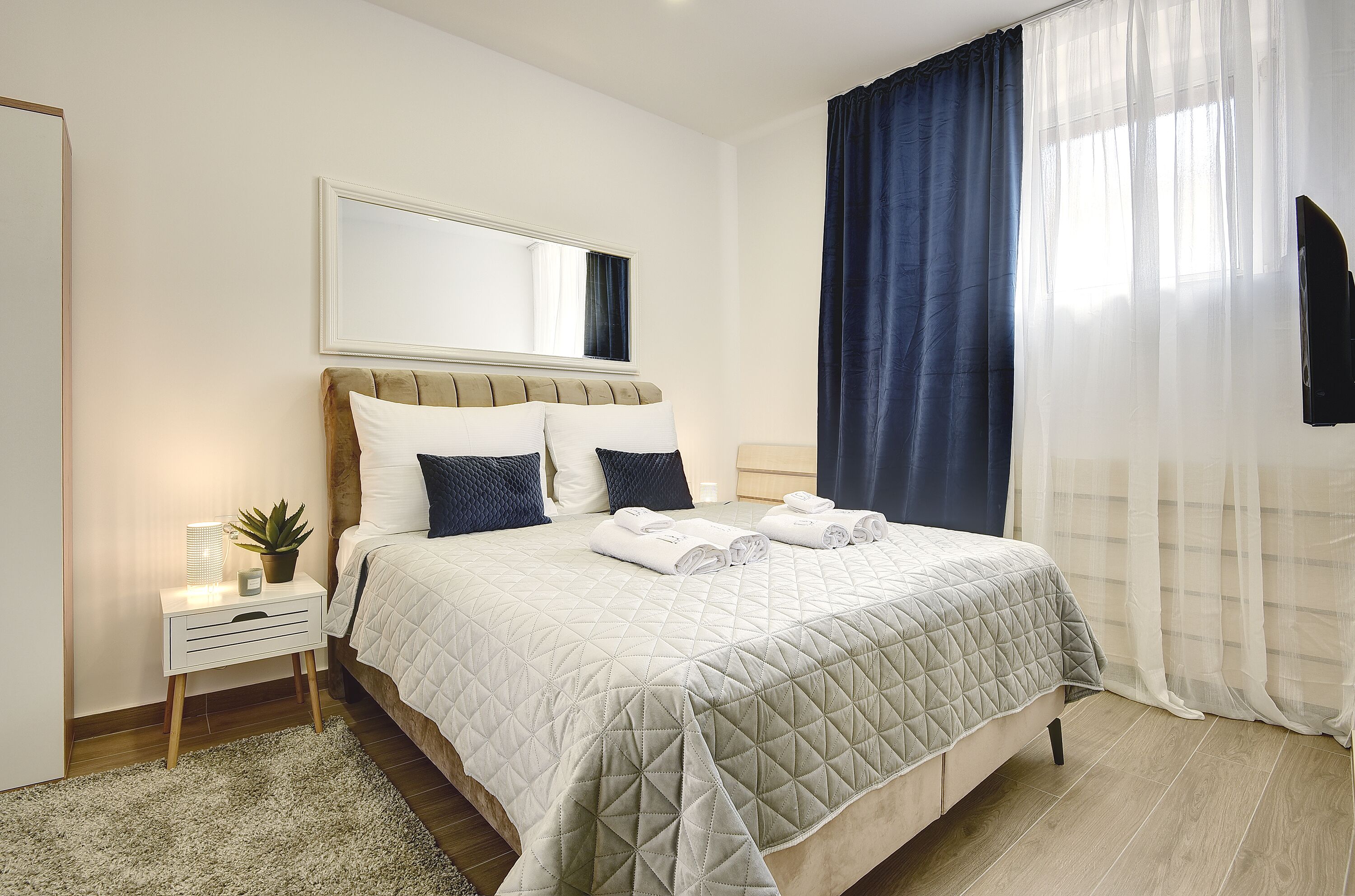 A neatly made bed with a quilted bedspread, a bedside table with a lamp and a plant, and a large mirror above the bed.