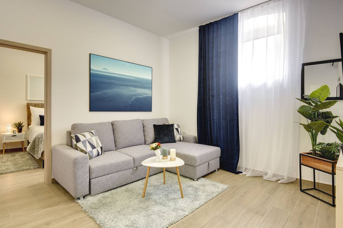 A modern living room with a grey sofa, a small white table with candles, and a large window with blue curtains.