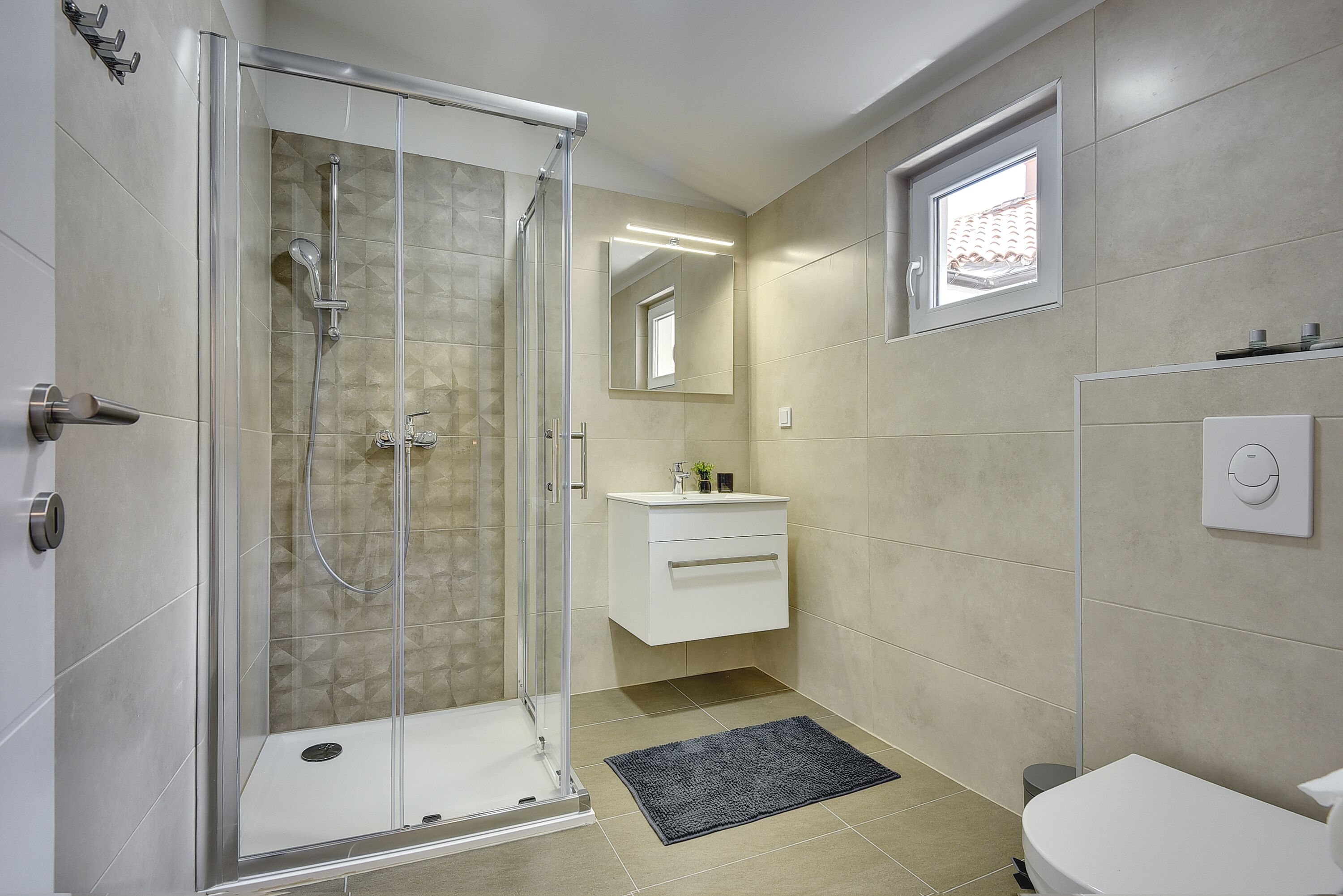 A modern bathroom with a glass-enclosed shower, a white vanity, and a small window.
