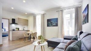 Una sala de estar moderna con un sofá gris, un comedor con mesa y sillas, y una cocina con armarios de madera y un extractor de aire negro.