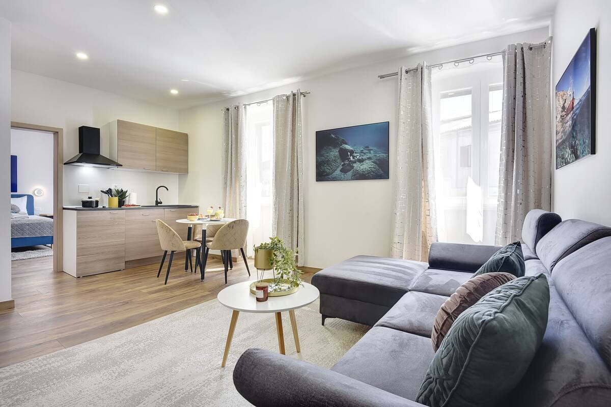A modern living room with a grey sofa, a dining area with a table and chairs, and a kitchen with wooden cabinets and a black range hood.