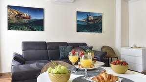 Una sala moderna con un sofá gris, una mesa de comedor con alimentos para el desayuno y dos cuadros enmarcados con motivos costeros.