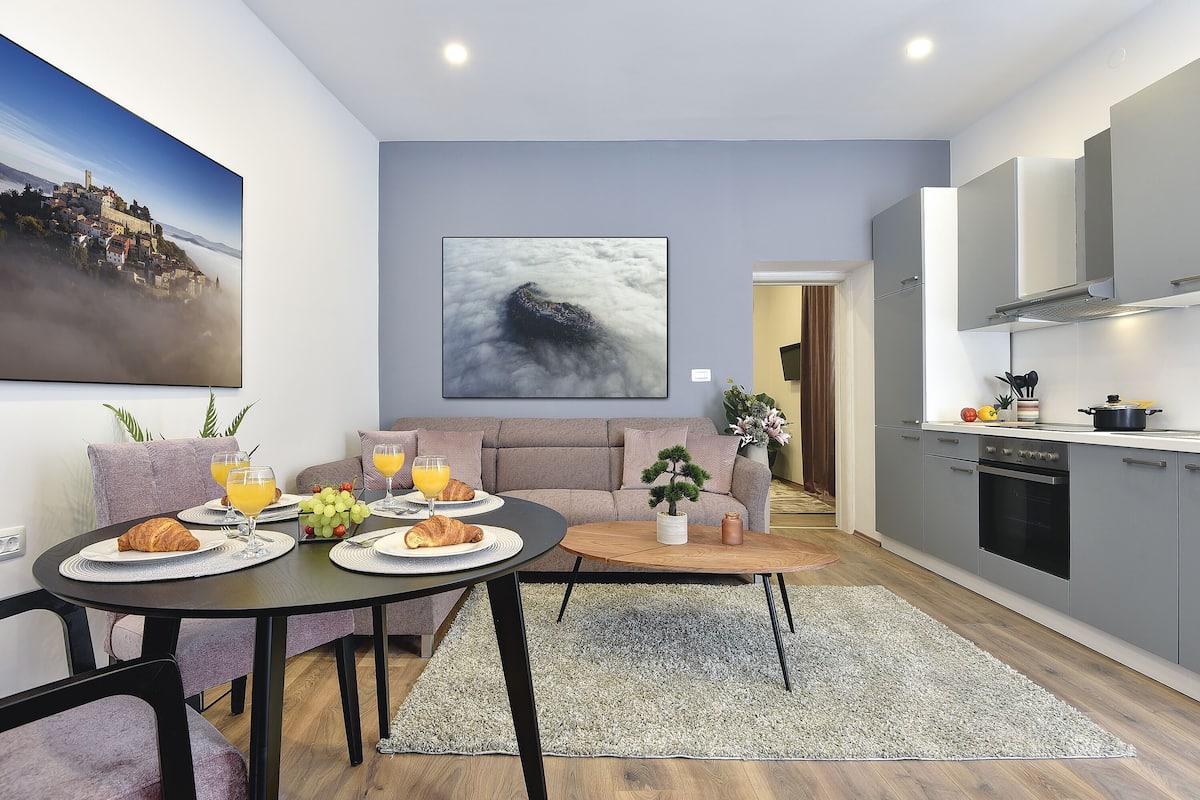 A modern kitchen with grey cabinets, a dining area with a round table, and a living room with a sofa.