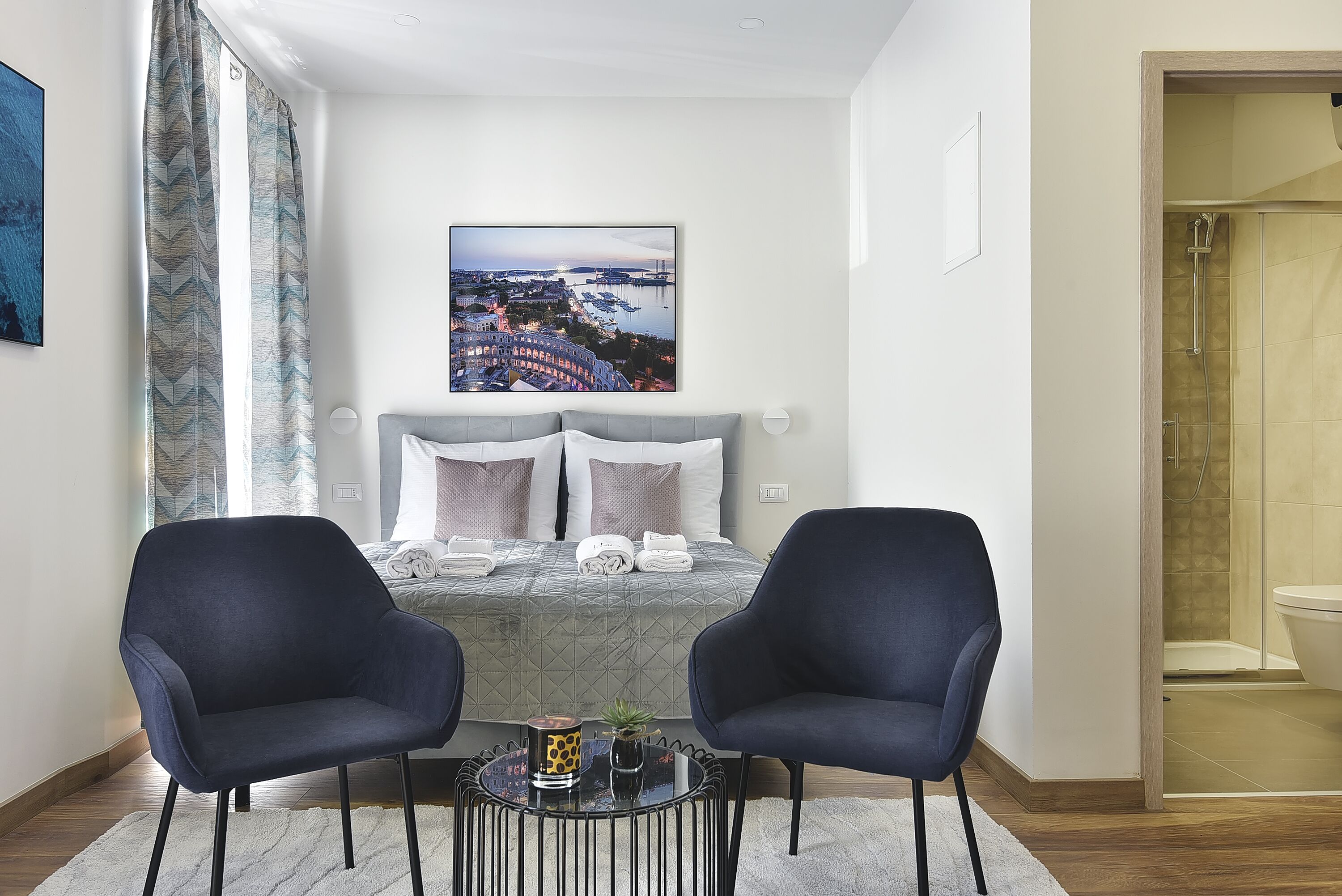 Un dormitorio de hotel moderno con una cama, dos sillones, una mesita con una planta y vistas a la ciudad.
