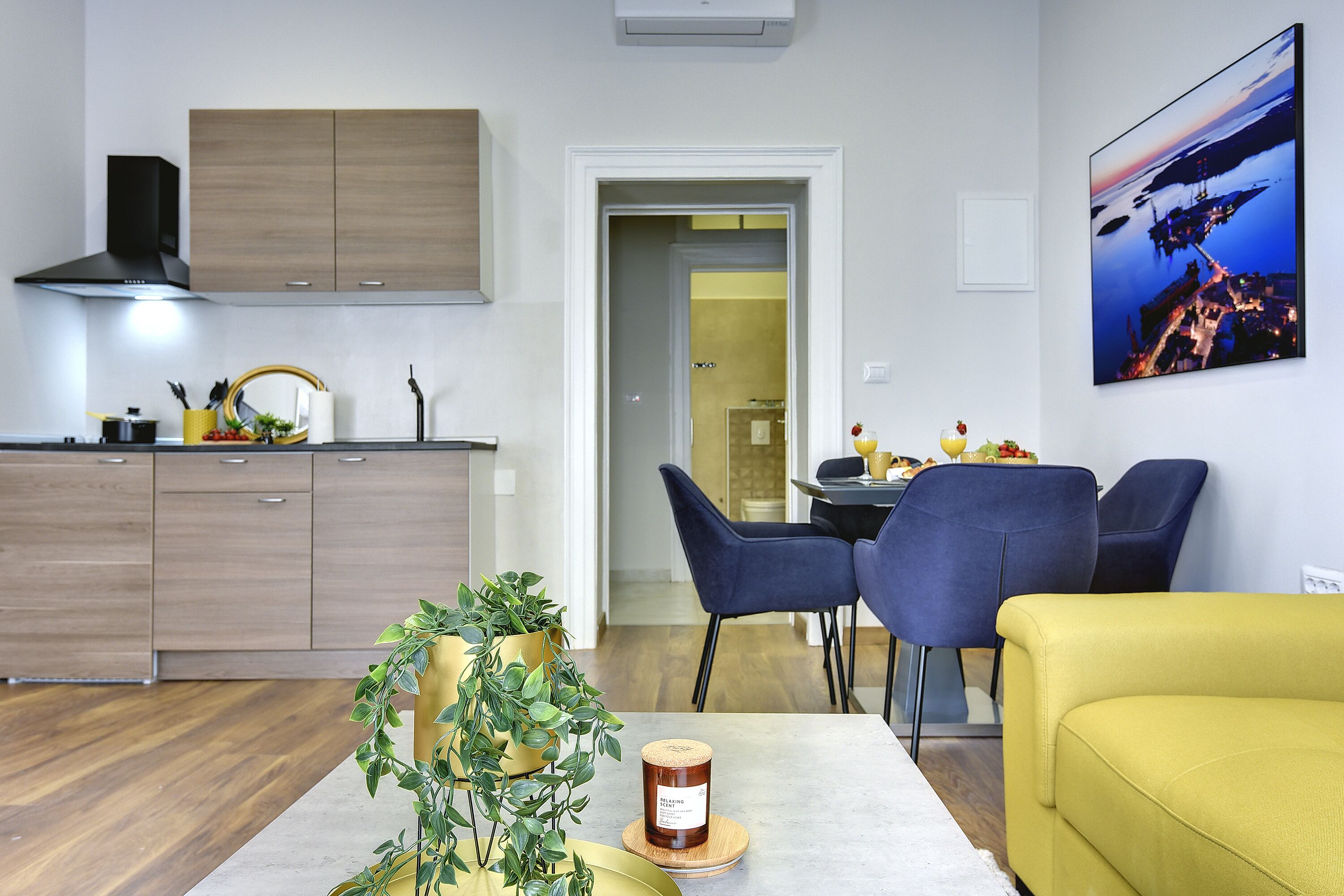 A modern kitchen with wooden cabinets, a dining area with blue chairs, and a yellow sofa.