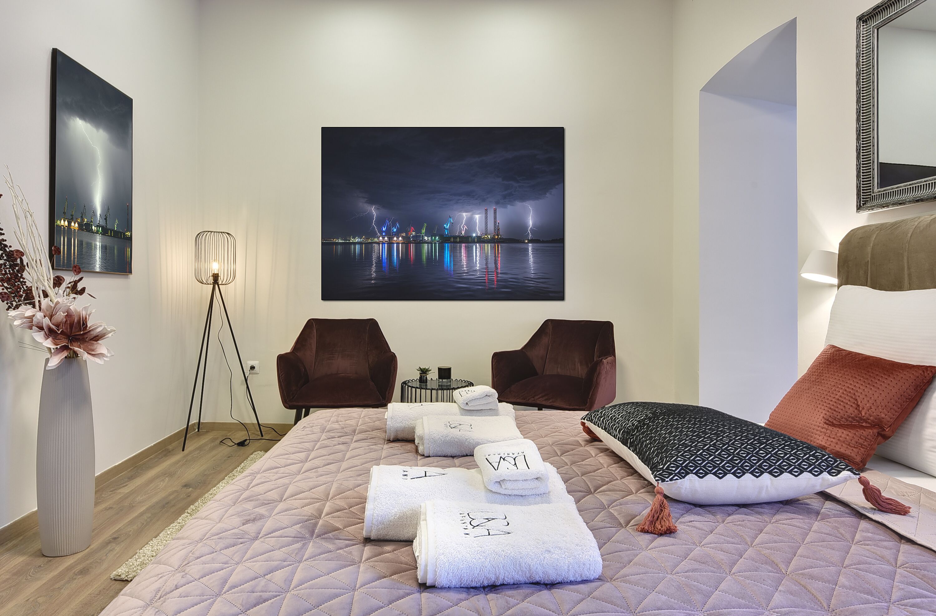Une chambre d’hôtel avec un lit, deux fauteuils, un grand tableau encadré représentant une vue de la ville et un vase avec des fleurs.