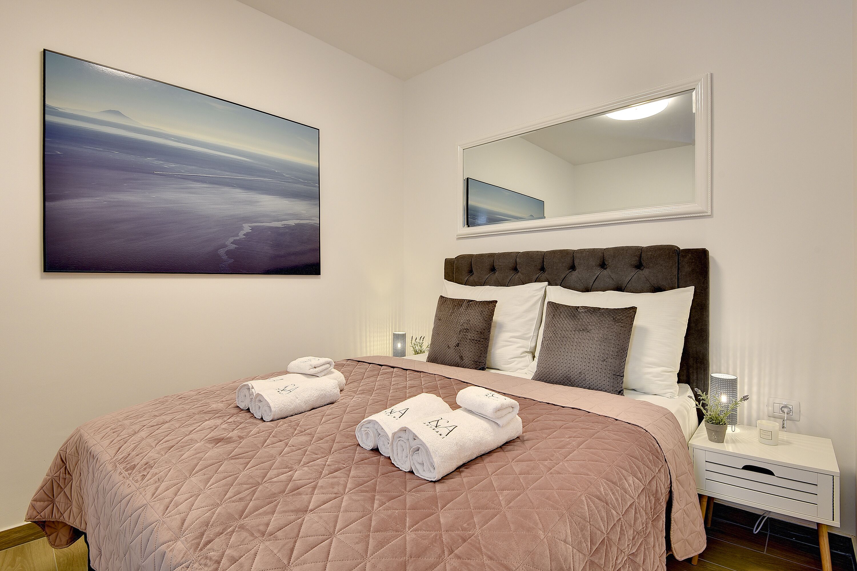 A neatly made bed with a quilted pink bedspread, two rolled towels, and a large landscape painting on the wall.