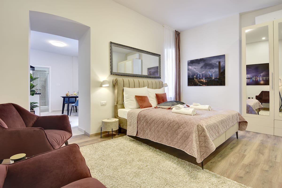 A modern bedroom with a large bed, a sitting area with a sofa, a small side table, and a painting on the wall.