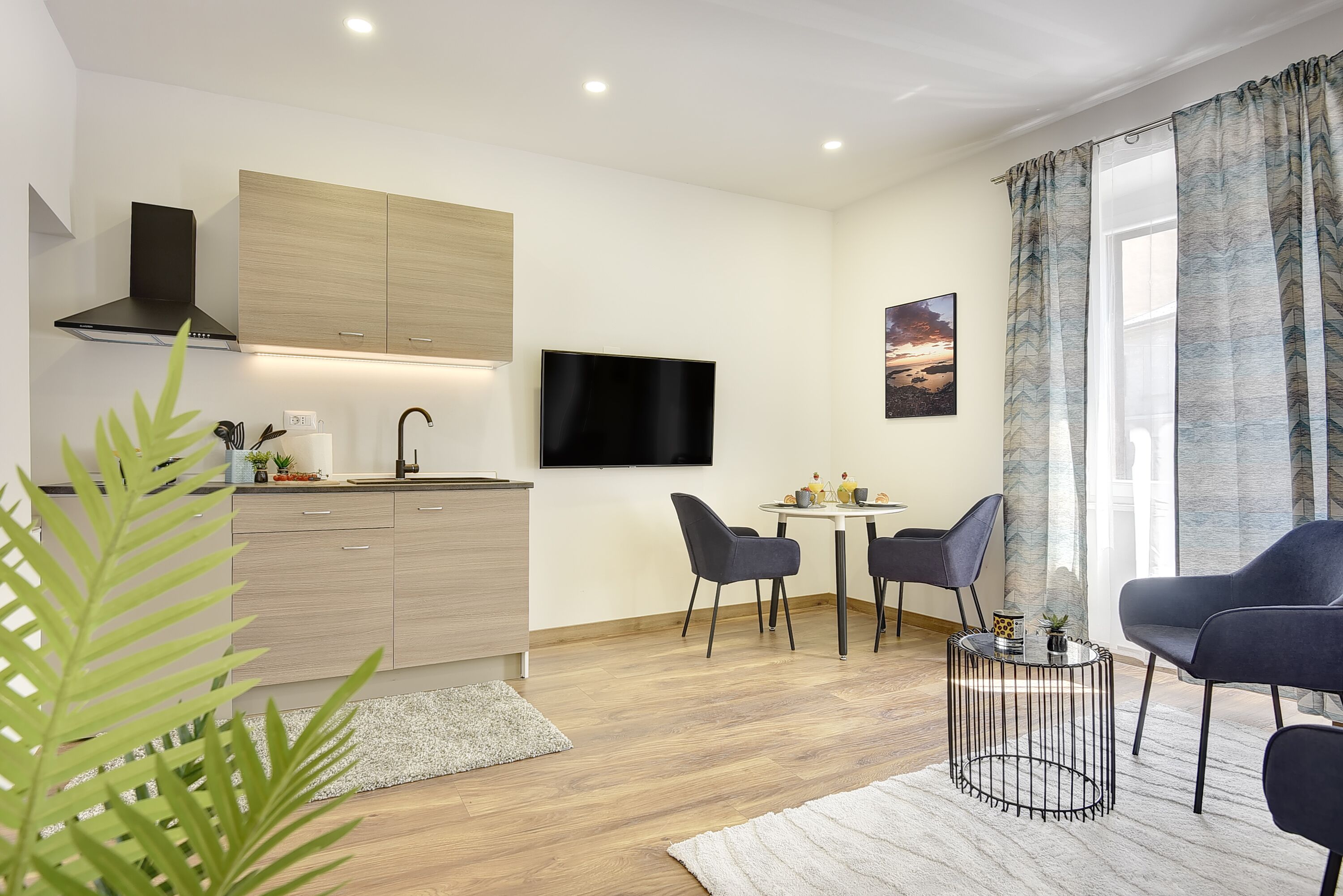 A modern living room with a dining area, a flat-screen TV mounted on the wall, and a large window with curtains.