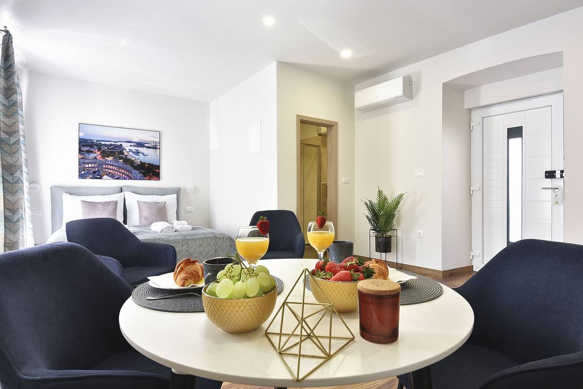 A modern hotel room with a round dining table, a sofa, a bed, and a view of a cityscape.