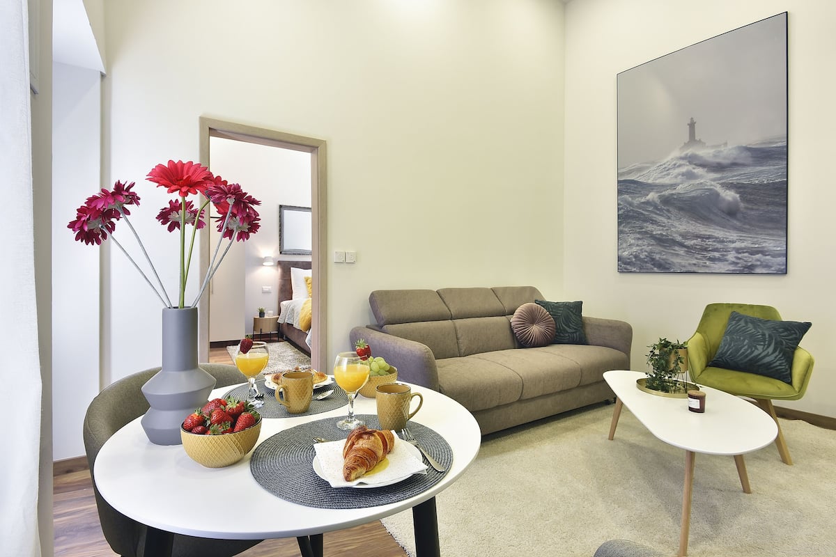 A living room with a dining table set for breakfast, a sofa, a green armchair, and a painting of a lighthouse on the wall.