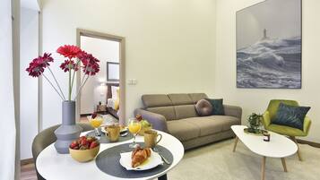 Una sala de estar con una mesa de comedor lista para el desayuno, un sofá, un sillón verde y un cuadro de un faro en la pared.
