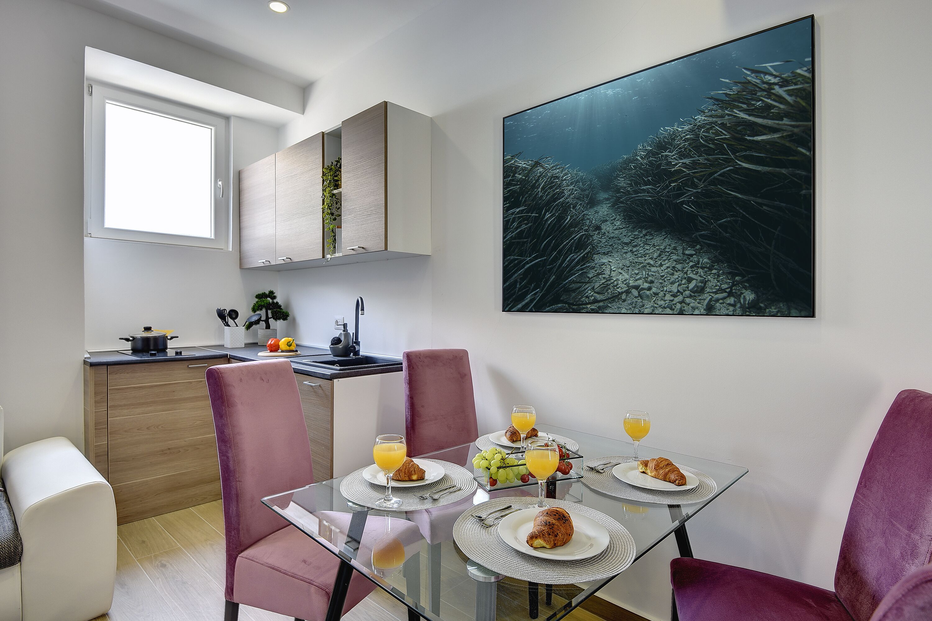 A modern kitchen with a dining area. The kitchen has wooden cabinets, a window, and a sink. The dining area has a glass table with plates, glasses, and food. There is a large painting on the wall.