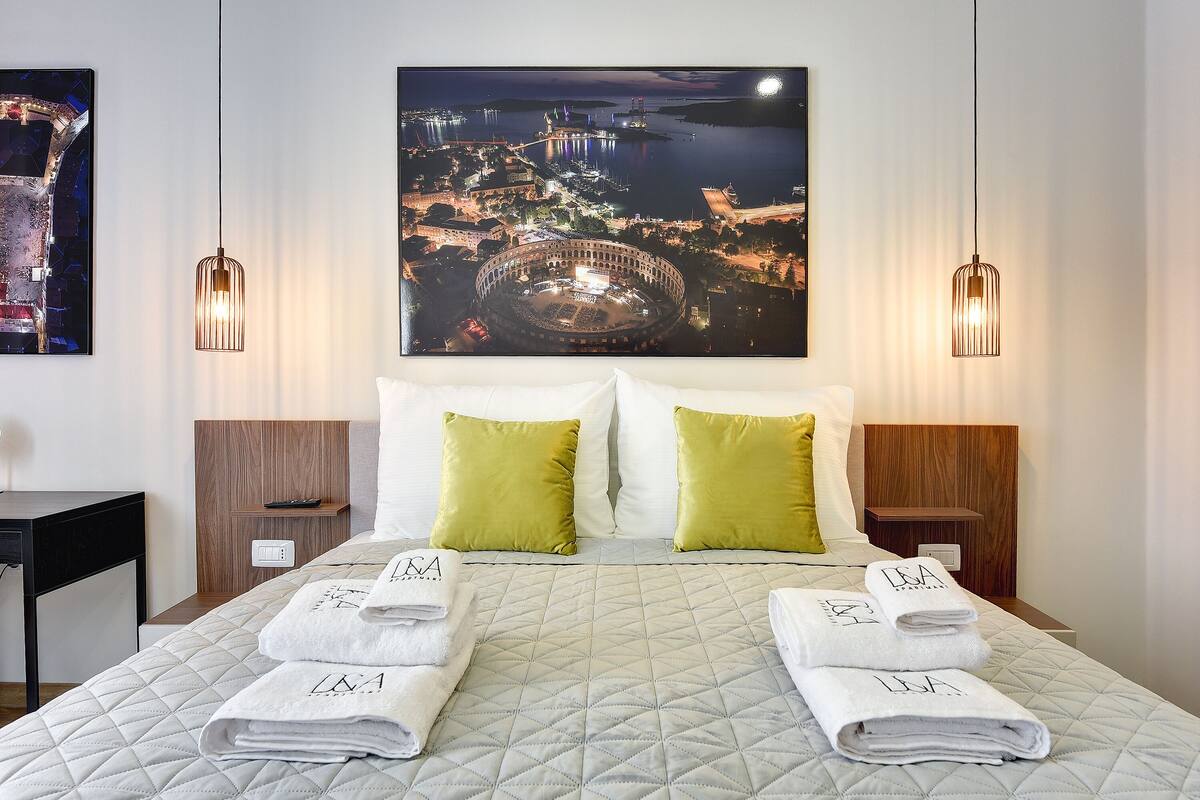 A hotel room with a bed, two green pillows, a nightstand, a desk, and a large framed picture of a cityscape at night.