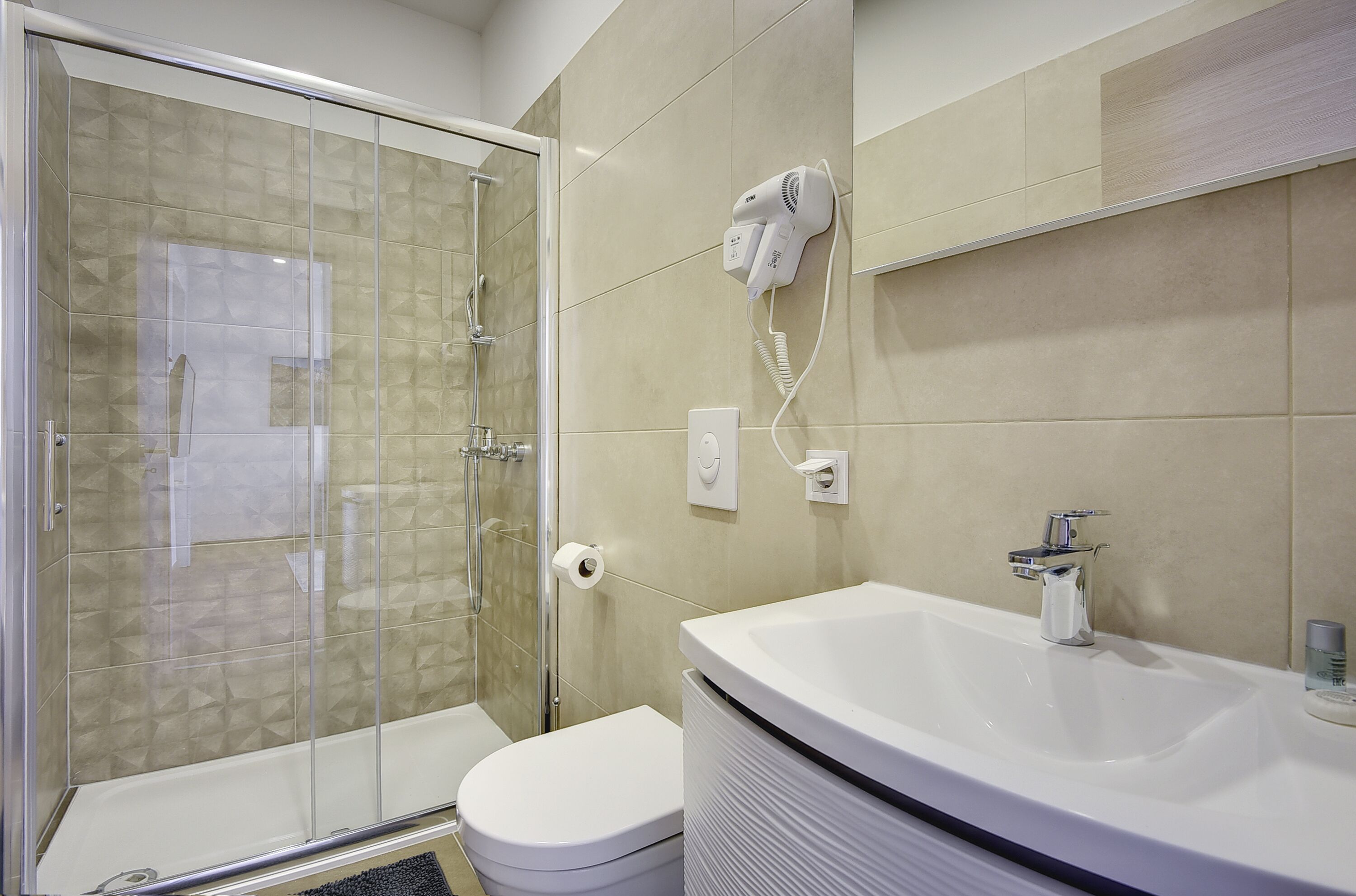 A bathroom with a glass shower enclosure, a white toilet, a sink with a mirror, and a hairdryer mounted on the wall.