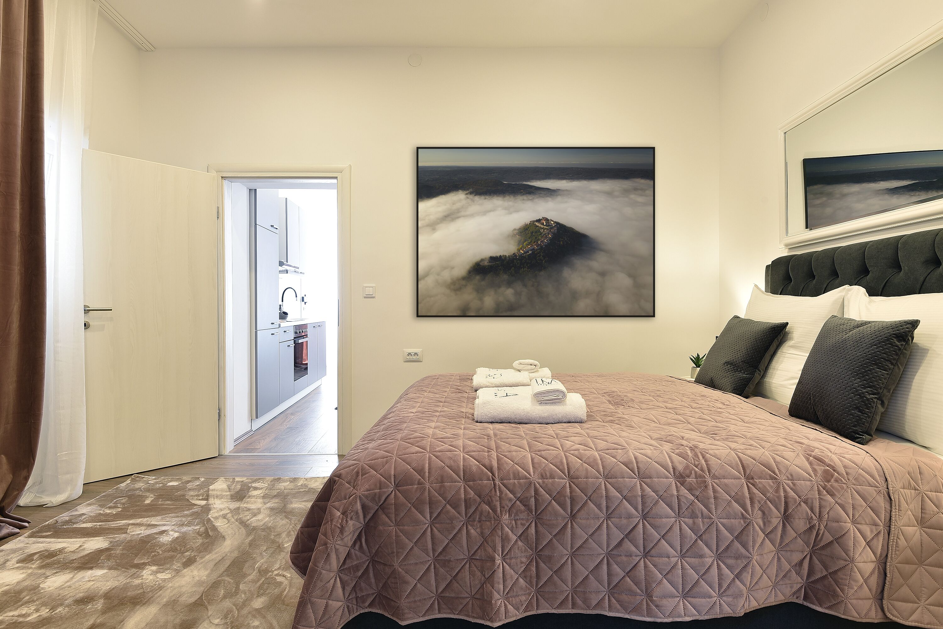 Un dormitorio con una cama grande, un cuadro de un paisaje, y vistas a una cocina y un baño a través de una puerta abierta.